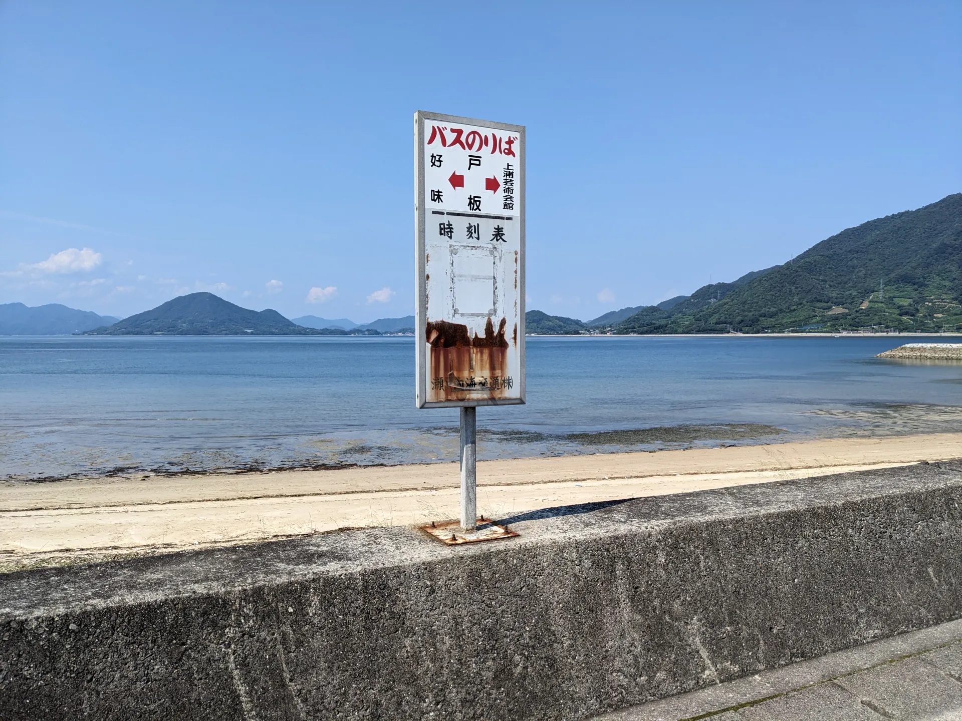 A rural Japanese bus stop sign reading バスのりば and 時刻表 beside the sea